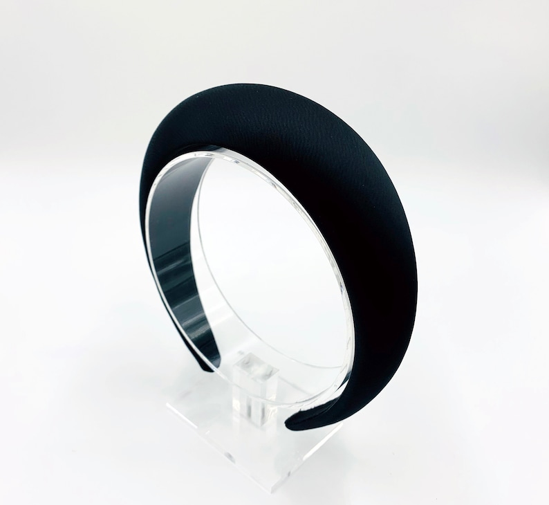 K&ouml;nnte beinhalten: Ein schwarzes Samt-Haarband mit einer breiten, abgerundeten Form. Das Haarband wird auf einem klaren Acryl-St&auml;nder pr&auml;sentiert.