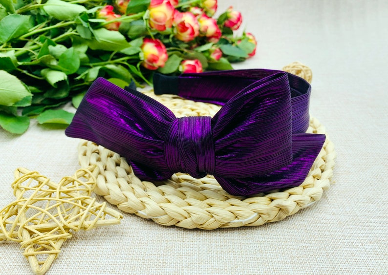 Pu&ograve; includere: Un cerchietto metallico viola con un grande fiocco. Il cerchietto &egrave; su una superficie di paglia intrecciata.