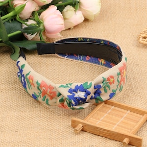 May include: A beige headband with a knotted design and floral embroidery in shades of blue, pink, and green.