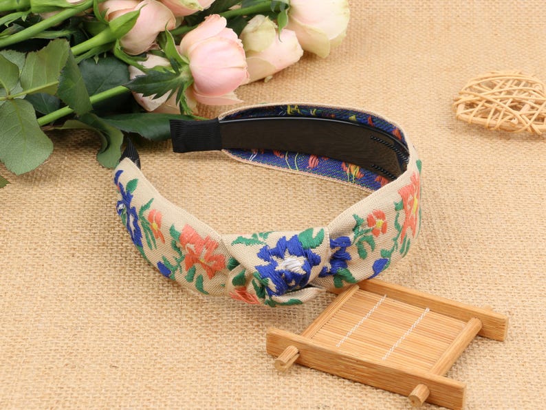 May include: A beige headband with a knotted design and floral embroidery in shades of blue, pink, and green.
