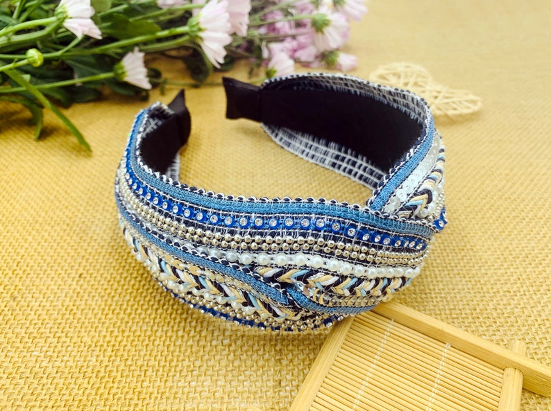 May include: A blue and white beaded headband with a knotted design. The headband is made of fabric and has a black elastic band.