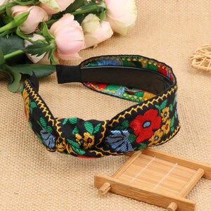 May include: A black headband with a knotted design and colorful floral embroidery. The headband features red, yellow, blue, and green flowers on a black background.