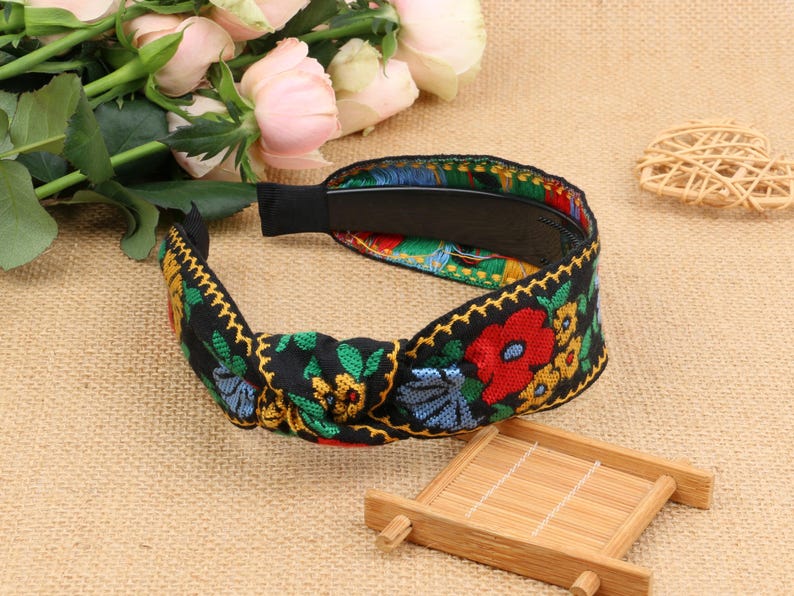 May include: A black headband with a knotted design and colorful floral embroidery. The headband features red, yellow, blue, and green flowers on a black background.
