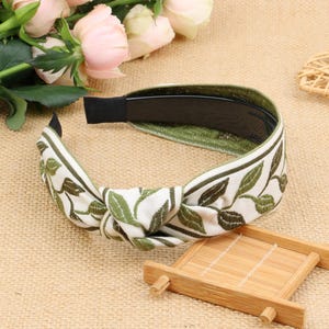 May include: A white headband with a green leaf pattern and a black elastic band. The headband is tied in a knot at the top.