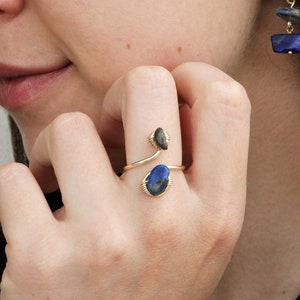 Könnte beinhalten: Ein goldener Ring mit einem blauen und grünen Stein, der in Draht gefasst ist. Der Ring wird am Zeigefinger einer Hand getragen.