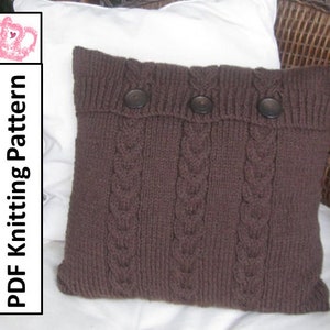 May include: A brown knitted pillow with a cable knit pattern and three brown buttons. The pillow is sitting on a white surface.