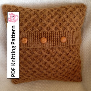 以下が含まれることがあります： 茶色の編み地のクッションで、織り模様と茶色のボタンが3つ付いています。クッションは白い表面の上に置かれています。PDF編み図です。