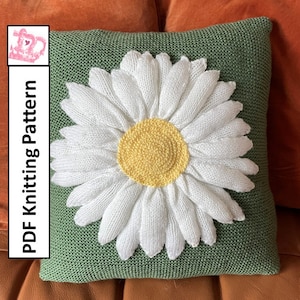 Puede incluir: Una almohada de punto verde con una flor de margarita blanca en el centro. La margarita tiene un centro amarillo y pétalos blancos.