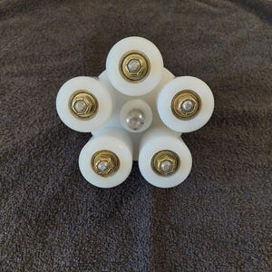 May include: A white, star-shaped object with five rounded wheels. Each wheel has a gold-coloured centre and a silver bolt. The central hub is silver. The object is on a grey, textured surface.