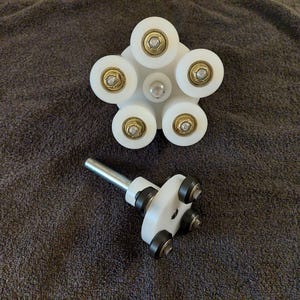 Op de afbeelding: Een witte kunststof rollerset met zes rollers en een metalen staaf. De rollers zijn in een cirkel gerangschikt en hebben een kleine metalen pin in het midden. De set staat op een grijs oppervlak.