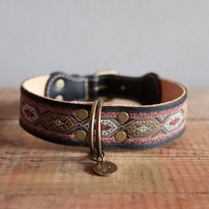Op de afbeelding: Een zwarte leren hondenriem met een rood, goud en wit gepatroneerd stoffen band. De riem heeft messing klinknagels en een messing D-ring. Een messing tag met een ontwerp is bevestigd aan de D-ring.