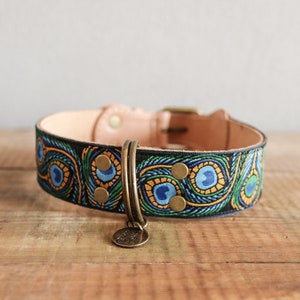 Op de afbeelding: Een hondenhalsband met een pauwenveermotief. De halsband heeft een zwarte basis met geborduurde blauwe, groene en gouden details. Het heeft een lichtbruin lederen binnenkant en een messing gesp en hardware. Er hangt een kleine, ronde bedel aan.
