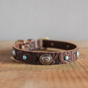 Può includere: Un collare per cani in pelle marrone con un design a rilievo floreale e dettagli argentati. Il collare ha pietre turchesi incastonate nel design.