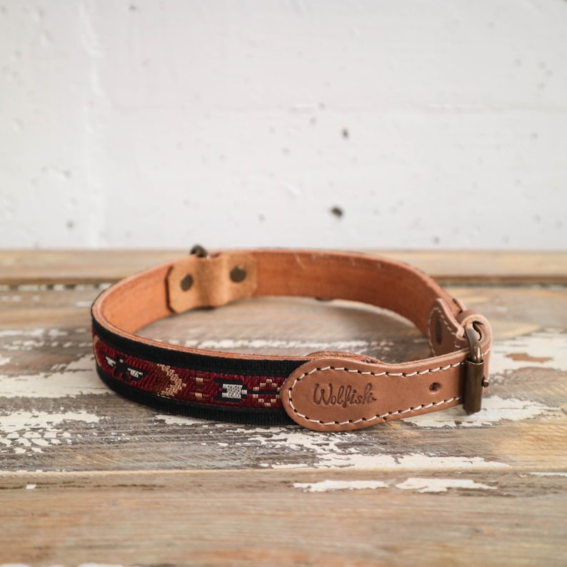 May include: A brown leather dog collar with a black and red patterned fabric insert. The collar has a brass buckle and the word "Wolfish" is embossed on the leather.
