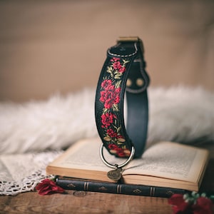 K&ouml;nnte beinhalten: Ein schwarzes Lederhundekragen mit roter und goldener Blumenstickerei. Das Halsband hat einen D-Ring aus Messing und ein kleines Messing-Schild mit der Gravur "Wanderlust".