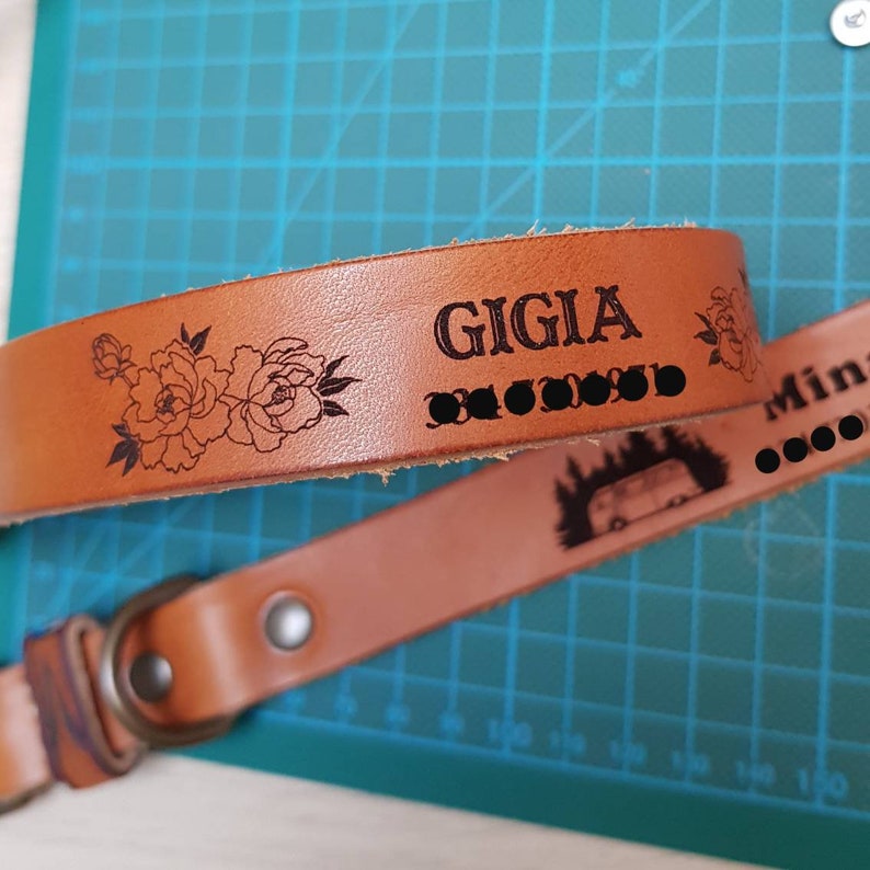 Puede incluir: Una correa de c&aacute;mara de cuero marr&oacute;n con el nombre "GIGIA" y un dise&ntilde;o floral grabado. La correa tambi&eacute;n tiene una hebilla de metal.