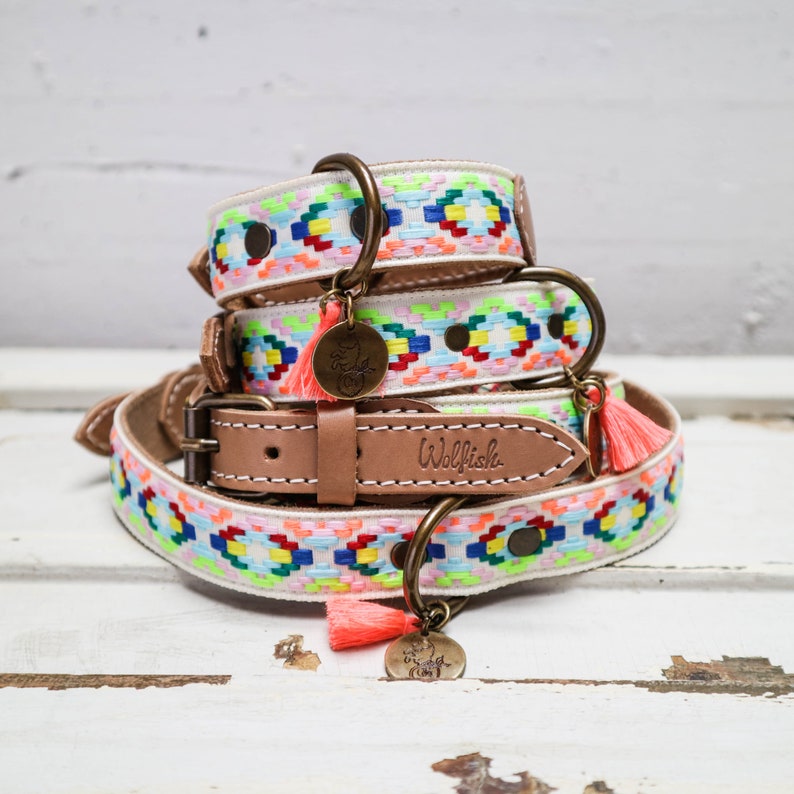 May include: A stack of three colorful dog collars with a white background and a pink tassel. The collars are decorated with a geometric pattern and the top collar has a brass tag that says "Wolfish".