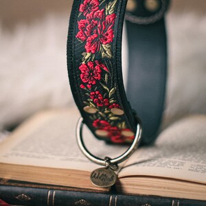 K&ouml;nnte beinhalten: Ein schwarzes Lederhundekragen mit einem roten und goldenen floralen Stickmuster. Das Halsband hat einen silbernen D-Ring und ein silbernes Schild mit der Aufschrift "Walk This Way".