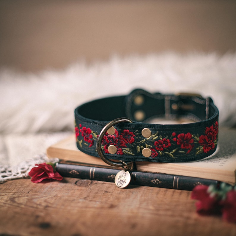 K&ouml;nnte beinhalten: Schwarzes Hundehalsband mit roten gestickten Blumen und einem goldenen Metalletikett. Das Halsband liegt auf einem Stapel B&uuml;cher.