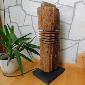 Könnte beinhalten: Eine hohe Holzlampe mit rustikalem, natürlichem Finish. Der Lampenfuß besteht aus übereinander gestapelten Holzstücken und hat einen schwarzen Metallfuß. Die Lampe ist beleuchtet und strahlt ein warmes Licht aus.