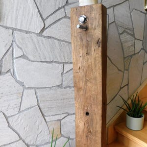 Könnte beinhalten: Ein hoher, rechteckiger, verwitterter brauner Holzpfosten mit einer glatten, hellen runden Oberseite. Eine kleine, dunkel bronzefarbene Figur einer Person steht oben auf. Ein silberner runder Knopf und ein kleiner Metallhaken sind an der Vorderseite angebracht.