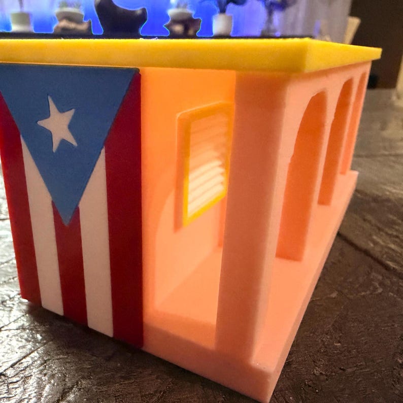 Pu&ograve; includere: Un modello architettonico stampato in 3D di un edificio con facciata color pesca, aperture ad arco e tetto giallo. Il lato della struttura presenta un design verticale della bandiera di Porto Rico con un triangolo blu, una stella bianca e strisce rosse.