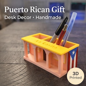 Pu&ograve; includere: Un organizer da scrivania stampato in 3D a forma di casa con tetto giallo, pareti rosa e la bandiera di Porto Rico sul lato. La parte superiore presenta tre scomparti contenenti un pennarello nero, una penna rossa e una blu. Il testo La Casita &egrave; in rilievo.