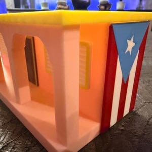 Pu&ograve; includere: Un modello stampato in 3D di una casa con pareti color pesca, un tetto giallo brillante e un portico con colonne ad arco. Il lato della struttura presenta un design verticale della bandiera di Porto Rico con un triangolo blu, una stella bianca e strisce rosse e bianche.