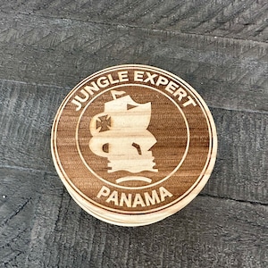 May include: A wooden coaster with a brown background and a white design. The design features a ship with a cross on the sail, the words "Jungle Expert" and "Panama".