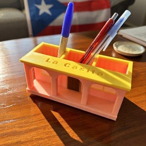 Pu&ograve; includere: Un organizer da scrivania rosa chiaro e giallo a forma di piccola casa con la scritta La Casita in rilievo sul tetto. L'organizer presenta tre scomparti rettangolari contenenti una penna blu, una penna rossa e una penna trasparente.