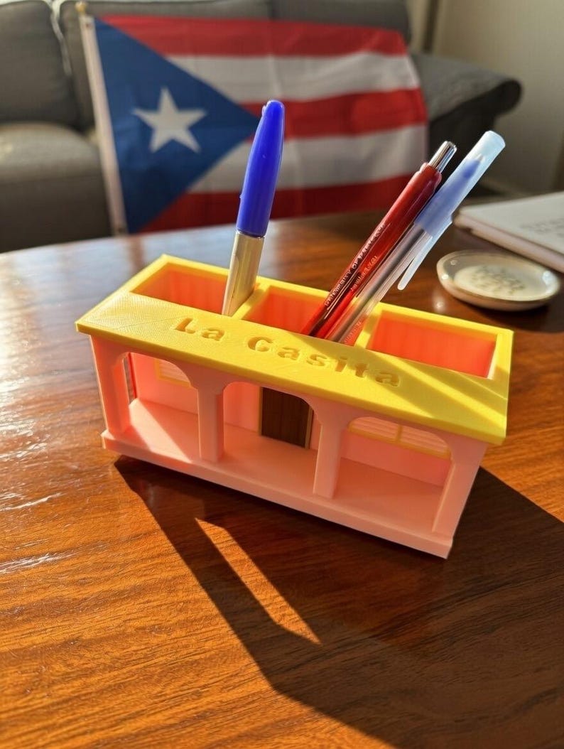 Pu&ograve; includere: Un organizer da scrivania rosa chiaro e giallo a forma di piccola casa con la scritta La Casita in rilievo sul tetto. L'organizer presenta tre scomparti rettangolari contenenti una penna blu, una penna rossa e una penna trasparente.