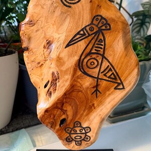 May include: Wooden art piece featuring a bird and spiral design, mounted on a black metal stand. The natural wood grain is visible, with a dark bird illustration and a turtle symbol at the bottom. A unique, handcrafted decorative item.