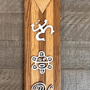 May include: A wooden plaque with a brown finish, featuring white relief carvings of Ta&iacute;no symbols. A triangle with a star is at the top. The plaque is rectangular and has a vertical orientation.