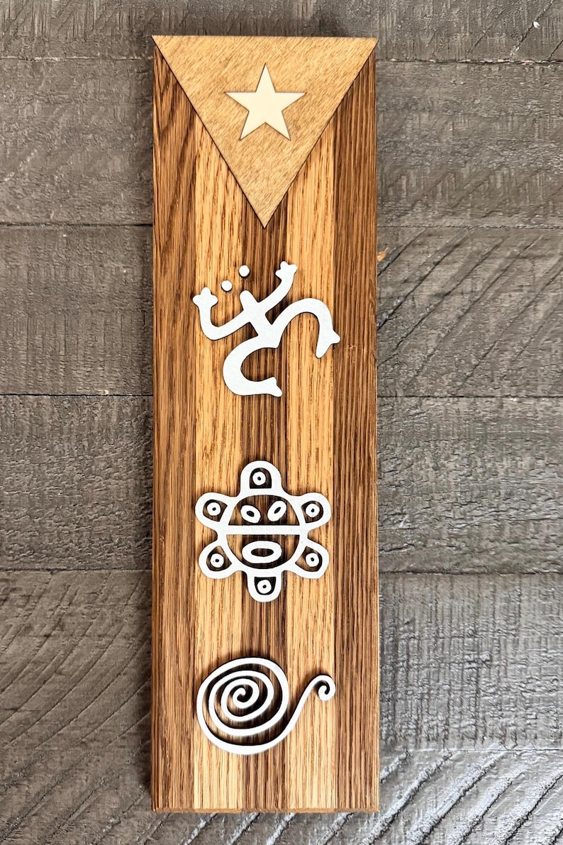 May include: A wooden plaque with a brown finish, featuring white relief carvings of Ta&iacute;no symbols. A triangle with a star is at the top. The plaque is rectangular and has a vertical orientation.