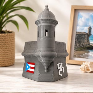 May include: A gray 3D-printed model of a garita watchtower featuring a small rectangular Puerto Rican flag emblem on the left side and a white Taino coqui symbol on the right side. The structure has a central turret with a dome top.