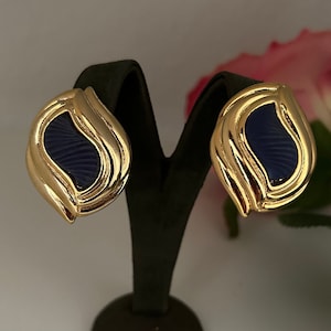 Puede incluir: Pendientes de clip dorados con un centro de esmalte azul marino. Los pendientes tienen una forma orgánica en capas. Los pendientes se exhiben en un soporte negro. Una rosa rosa es visible en el fondo.