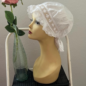 Op de afbeelding: Een witte muts met kanten rand en een decoratieve band, tentoongesteld op een mannequinhoofd. Een roze roos in een groene glazen fles staat in de buurt. De muts is in vintage stijl, mogelijk voor een kostuum of speciale gelegenheid.