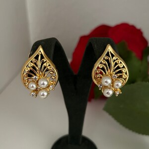 May include: A pair of gold earrings with a delicate filigree design and pearl accents. The earrings are displayed on a black stand.