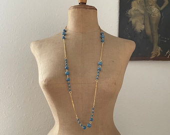 May include: A long necklace featuring alternating segments of gold-toned metal chain and round, light blue beads. The necklace is displayed on a beige fabric-covered mannequin torso, showcasing a repeating pattern of beads and textured metal links.