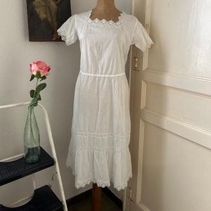 May include: A white cotton dress with lace trim. The dress has a square neckline, short sleeves, and a gathered skirt. The dress is on a white mannequin.