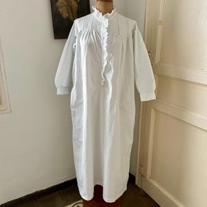 以下が含まれることがあります： 長袖の白いナイトガウン。アイレットレースのトリムが付いた高いフリル襟、胸元の縦プリーツ、そしてお揃いのフリルレースの縁取りで飾られた中央のボタンダウン前立てが特徴で、木製のドレスフォームにディスプレイされています。