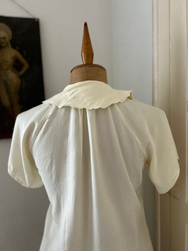 May include: A white, short-sleeved blouse with a scalloped collar. The blouse is on a white mannequin.