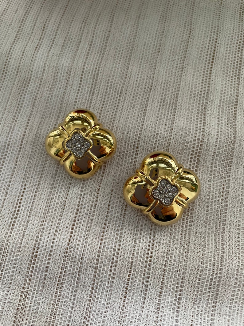 May include: A pair of gold tone earrings with a four-leaf clover design. Each clover has a center of clear rhinestones.