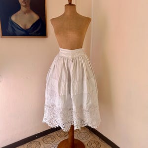 May include: A vintage white skirt with a detailed, eyelet lace hem is displayed on a wooden dress form. The skirt is gathered at the waist and falls to mid-calf. A framed portrait hangs on the wall in the background.