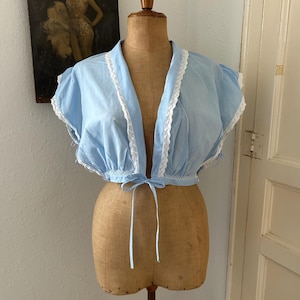 Peut inclure: Un chemisier court bleu clair avec des manches courtes à volants et une bordure en dentelle blanche le long de l'ouverture avant et des manches. Le vêtement présente une taille froncée avec un lien de serrage en tissu bleu clair.