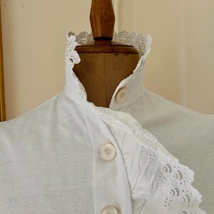 May include: Close-up of a white blouse with a high, ruffled collar and decorative lace trim. The blouse features a series of small, textured diamond patterns and large, round buttons. The garment is displayed on a wooden mannequin.