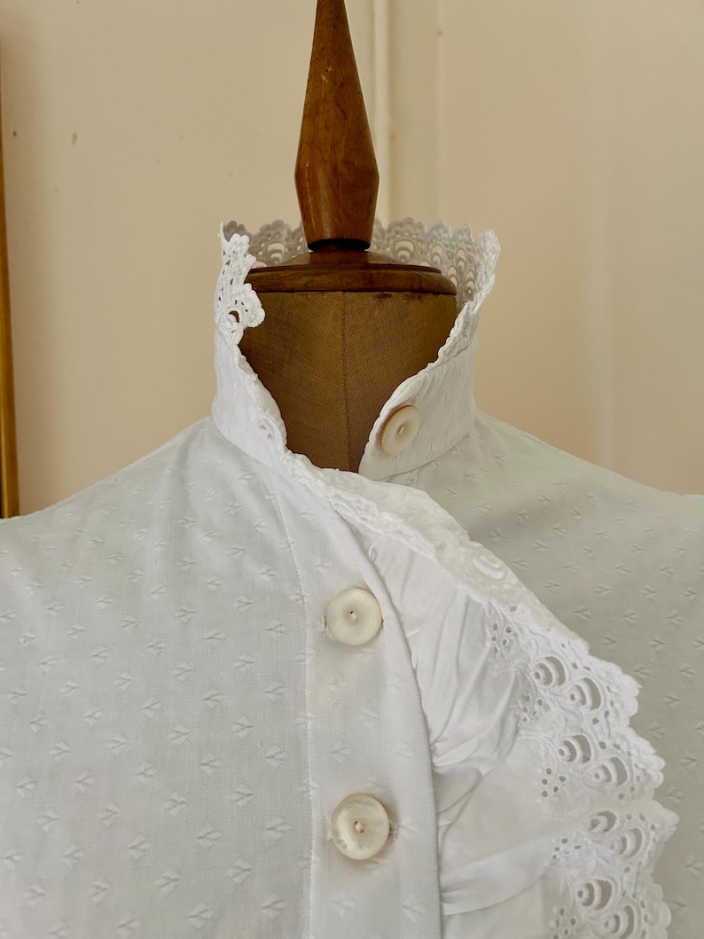 May include: Close-up of a white blouse with a high, ruffled collar and decorative lace trim. The blouse features a series of small, textured diamond patterns and large, round buttons. The garment is displayed on a wooden mannequin.