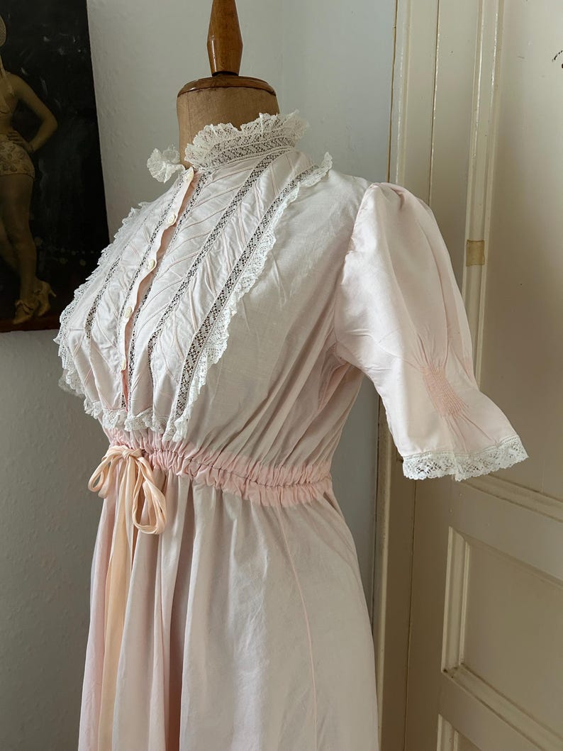 K&ouml;nnte beinhalten: Ein rosafarbenes Vintage-Nachthemd mit wei&szlig;en Spitzenbesatz und einer Schleife an der Taille.