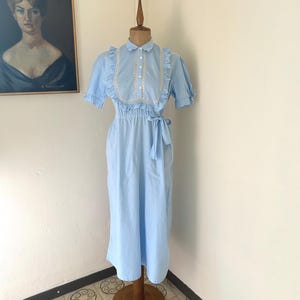 May include: A light blue vintage dress on a wooden mannequin. The dress has short sleeves, a collared neckline with white trim, and a waist tie. The floor-length dress has a simple design. The background features a vintage painting. The dress is approximately 140 cm in length.