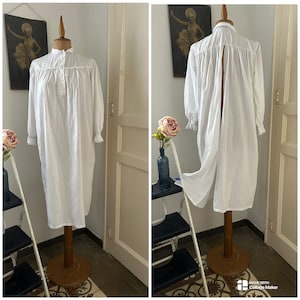 Könnte beinhalten: Ein weißes Nachthemd aus Baumwolle mit einem langen, fließenden Rock und einem geteilten Rücken. Das Nachthemd hat einen hohen Kragen und lange Ärmel mit Spitzenbesatz. Es wird auf einer Holzpuppe präsentiert.